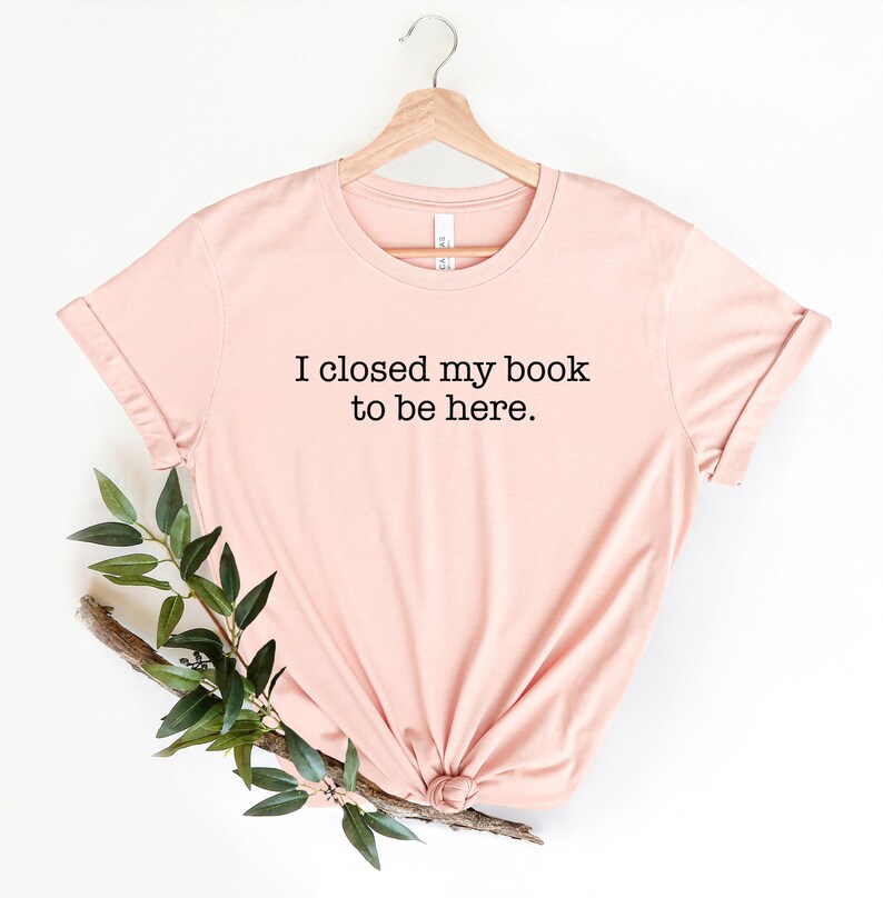 K&ouml;nnte beinhalten: Ein hellrosa T-Shirt mit dem Text "Ich habe mein Buch geschlossen, um hier zu sein." in Schwarz gedruckt.