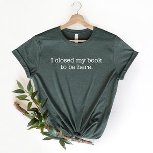 K&ouml;nnte beinhalten: Ein dunkelgr&uuml;nes T-Shirt mit dem Text "Ich habe mein Buch geschlossen, um hier zu sein." in wei&szlig; auf der Vorderseite.