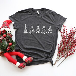 K&ouml;nnte beinhalten: Ein dunkelgraues T-Shirt mit einem wei&szlig;en Design aus f&uuml;nf stilisierten Weihnachtsb&auml;umen. Die B&auml;ume sind in einer Reihe &uuml;ber die Mitte des Shirts angeordnet.