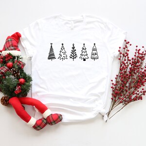 K&ouml;nnte beinhalten: Wei&szlig;es T-Shirt mit einer schwarz-wei&szlig;en Grafik von f&uuml;nf stilisierten Weihnachtsb&auml;umen.