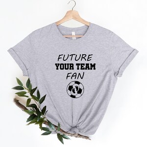 May include: Gray t-shirt with the text "FUTURE YOUR TEAM FAN" and a soccer ball with baby footprints on it.