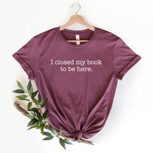 K&ouml;nnte beinhalten: Ein maroonfarbenes T-Shirt mit dem Aufdruck "Ich habe mein Buch geschlossen, um hier zu sein." in wei&szlig;.