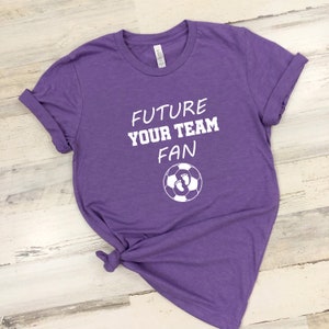 May include: Purple t-shirt with white text that reads "Future Your Team Fan" and a white soccer ball with two white footprints inside.
