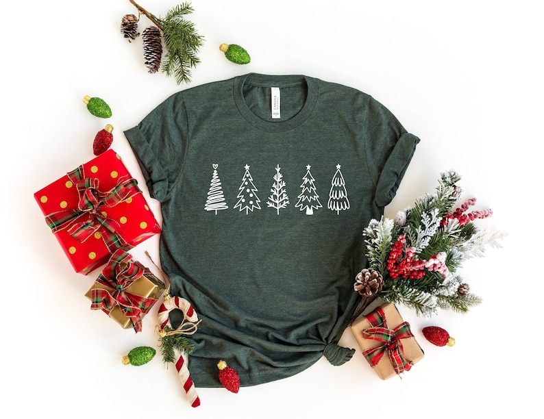 K&ouml;nnte beinhalten: Ein dunkelgr&uuml;nes T-Shirt mit einem wei&szlig;en Design aus f&uuml;nf stilisierten Weihnachtsb&auml;umen. Das Shirt ist umgeben von Weihnachtsdekorationen, darunter verpackte Geschenke, Kiefern&auml;ste und Ornamente.