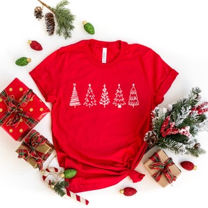 K&ouml;nnte beinhalten: Rotes T-Shirt mit einem wei&szlig;en Umriss von f&uuml;nf Weihnachtsb&auml;umen auf der Vorderseite. Das Shirt ist von Weihnachtsdekorationen umgeben, darunter verpackte Geschenke, Ornamente und ein Tannenzweig.