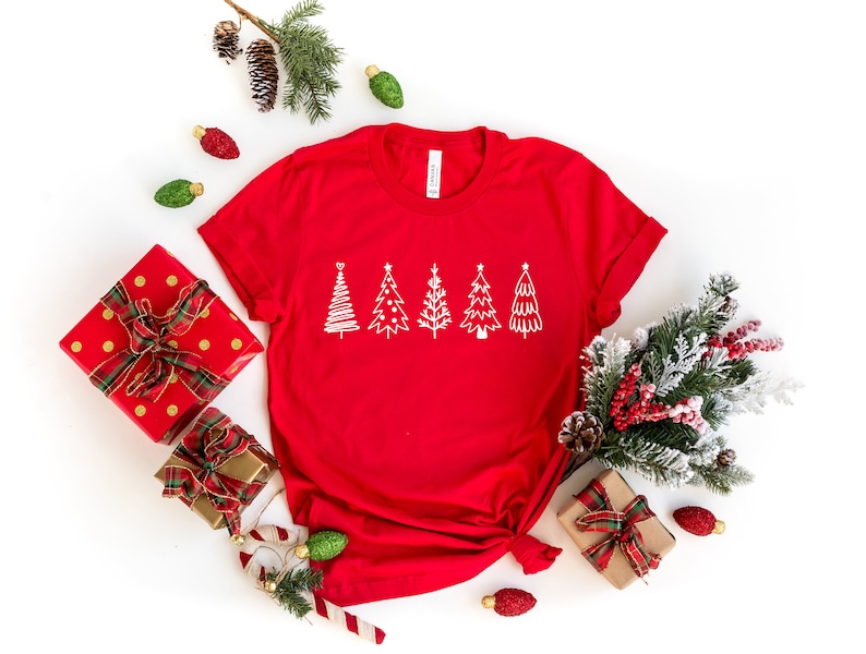 K&ouml;nnte beinhalten: Rotes T-Shirt mit einem wei&szlig;en Umriss von f&uuml;nf Weihnachtsb&auml;umen auf der Vorderseite. Das Shirt ist von Weihnachtsdekorationen umgeben, darunter verpackte Geschenke, Ornamente und ein Tannenzweig.