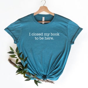 K&ouml;nnte beinhalten: T-Shirt in Tealblau mit wei&szlig;em Text, der "Ich habe mein Buch geschlossen, um hier zu sein." lautet.