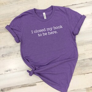 K&ouml;nnte beinhalten: Lilafarbenes T-Shirt mit wei&szlig;em Text, der "Ich habe mein Buch geschlossen, um hier zu sein." lautet.