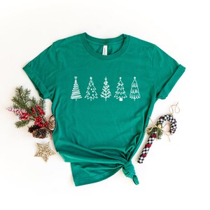 K&ouml;nnte beinhalten: Ein gr&uuml;nes T-Shirt mit einem wei&szlig;en Design aus f&uuml;nf stilisierten Weihnachtsb&auml;umen. Das Shirt ist unten verknotet und liegt auf einer wei&szlig;en Oberfl&auml;che.