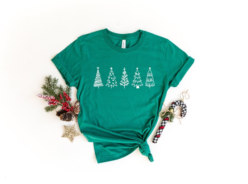 K&ouml;nnte beinhalten: Ein gr&uuml;nes T-Shirt mit einem wei&szlig;en Design aus f&uuml;nf stilisierten Weihnachtsb&auml;umen. Das Shirt ist unten verknotet und liegt auf einer wei&szlig;en Oberfl&auml;che.