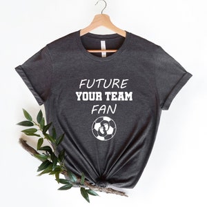 May include: A dark gray t-shirt with the text "FUTURE YOUR TEAM FAN" and a soccer ball with footprints on it.
