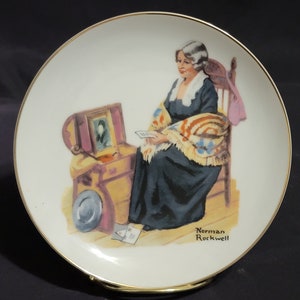 Peut inclure: Une assiette en céramique blanche avec un bord doré présente une peinture d'une femme assise sur une chaise, tenant une lettre. La femme porte une robe bleue et une couverture rouge, blanche et bleue. L'assiette présente également un coffre en bois avec un cadre photo sur le dessus. L'assiette est signée "Norman Rockwell" dans le coin inférieur droit.