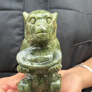 May include: A carved green stone monkey figurine holding a bowl. The monkey is in a seated position with its legs bent. The figurine has detailed facial features and is made of a speckled green stone.