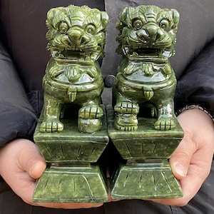 Può includere: Due statue di cani Foo verde giada. Ogni statua è scolpita nei dettagli, raffigurante una creatura simile a un leone con un'espressione feroce, seduta su una base quadrata. Le statue sono tenute davanti a uno sfondo scuro.
