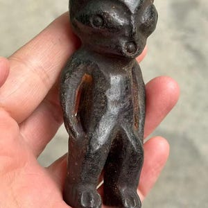 May include: A small, dark-colored figurine of a humanoid figure with large eyes and a small mouth. The figure has a simple, stylized form with arms at its sides and feet. The material appears to be a dark metal or stone, with a weathered appearance.