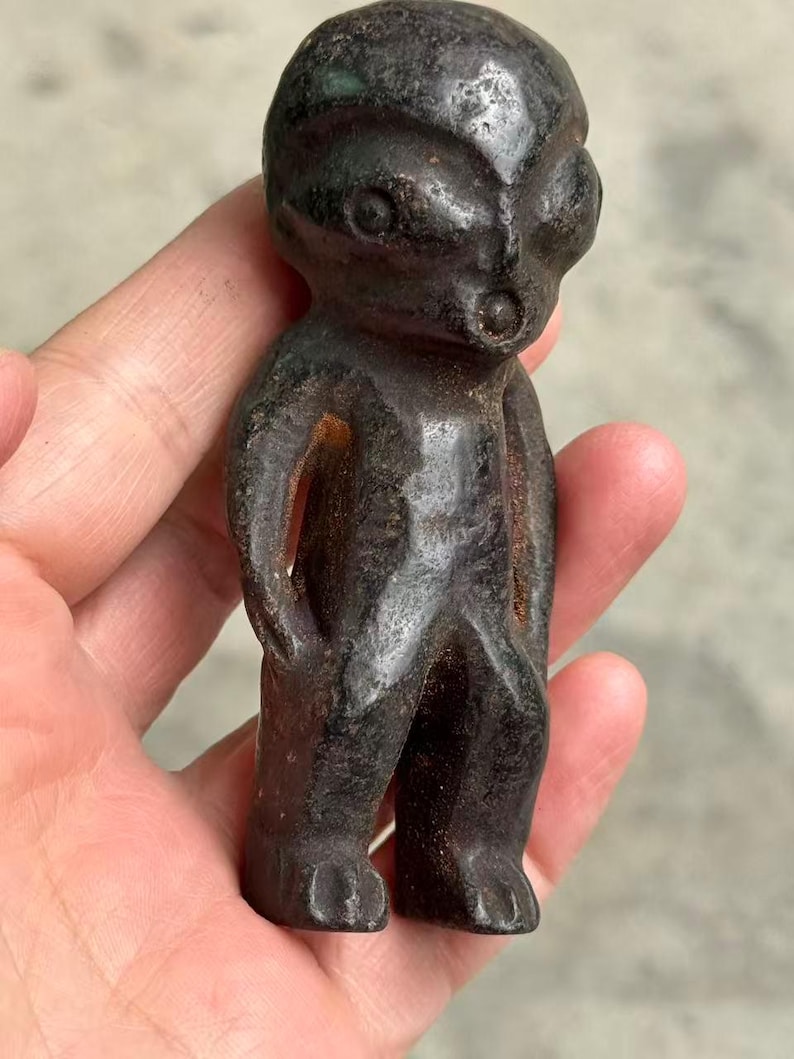 May include: A small, dark-colored figurine of a humanoid figure with large eyes and a small mouth. The figure has a simple, stylized form with arms at its sides and feet. The material appears to be a dark metal or stone, with a weathered appearance.