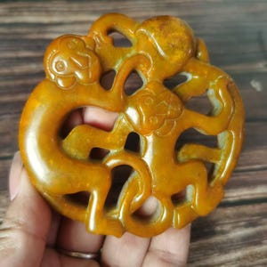 Może przedstawiać: Rzeźbiony, bursztynowy wisiorek z wystylizowanym wzorem dwóch splecionych postaci. Wisiorek ma gładką, wypolerowaną powierzchnię i skomplikowane detale. Postacie wydają się być zwierzętami lub mitycznymi stworzeniami.