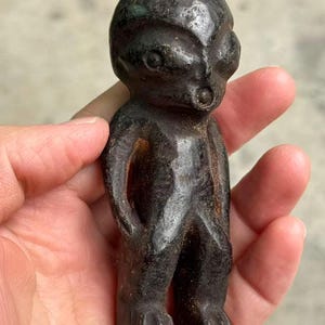 May include: A dark, weathered figurine of a humanoid alien with large eyes and a small mouth. The figure is standing with arms at its sides and feet planted. The sculpture has a rough, textured surface and is dark brown in color.