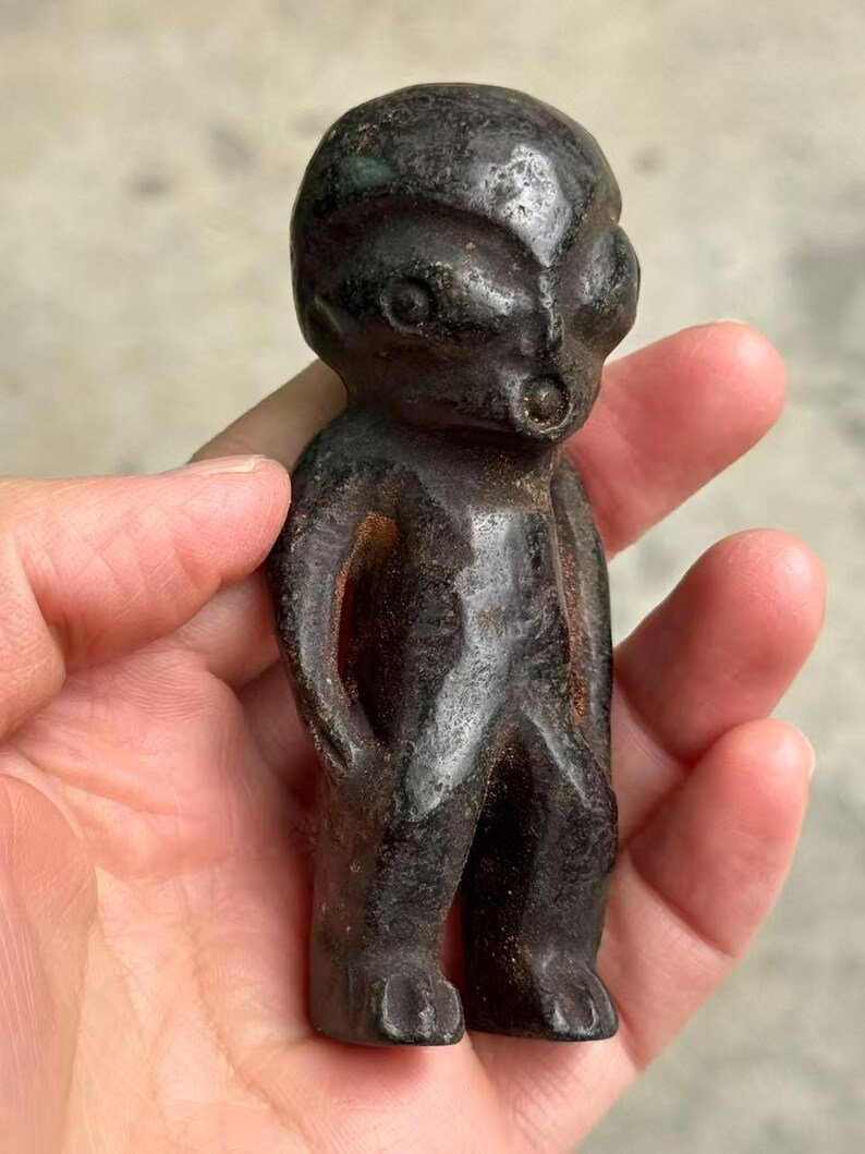 May include: A dark, weathered figurine of a humanoid alien with large eyes and a small mouth. The figure is standing with arms at its sides and feet planted. The sculpture has a rough, textured surface and is dark brown in color.