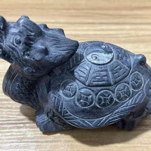 以下が含まれることがあります： 龍の頭を持つ亀の形をしたダークグレーの石の彫刻。甲羅には中央に陰陽のシンボルが刻まれ、その周囲を円形のコインのモチーフと幾何学模様が囲んでいます。龍の頭は横を向いています。