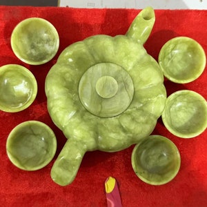 May include: A light green stone tea set consisting of a round, pumpkin-shaped teapot with a matching lid and six small, circular tea cups. The pieces feature a smooth, polished finish with natural marbled patterns throughout the light green stone.