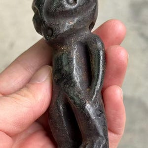 May include: A dark gray, carved figurine of a humanoid figure with a rounded head, large eye, and small arms held behind its back. The sculpture has a smooth, polished surface and is held in a hand.