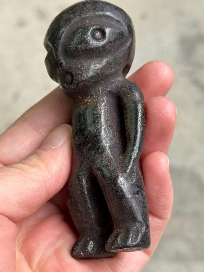 May include: A dark gray, carved figurine of a humanoid figure with a rounded head, large eye, and small arms held behind its back. The sculpture has a smooth, polished surface and is held in a hand.