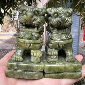 May include: Two jade lion statues, each carved with intricate details. The statues are a muted green color and sit on rectangular bases. The lions have fierce expressions and are posed in a seated position. The statues are likely decorative items.
