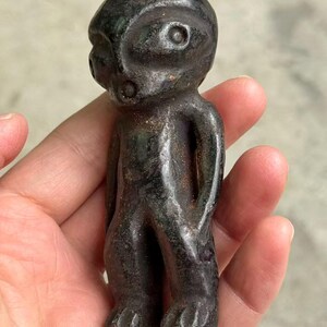 May include: A dark, weathered figurine of a humanoid figure with a large head and small body. The figure has a rounded helmet-like head with two eye-like indentations and a small mouth. The figure is held in a hand.