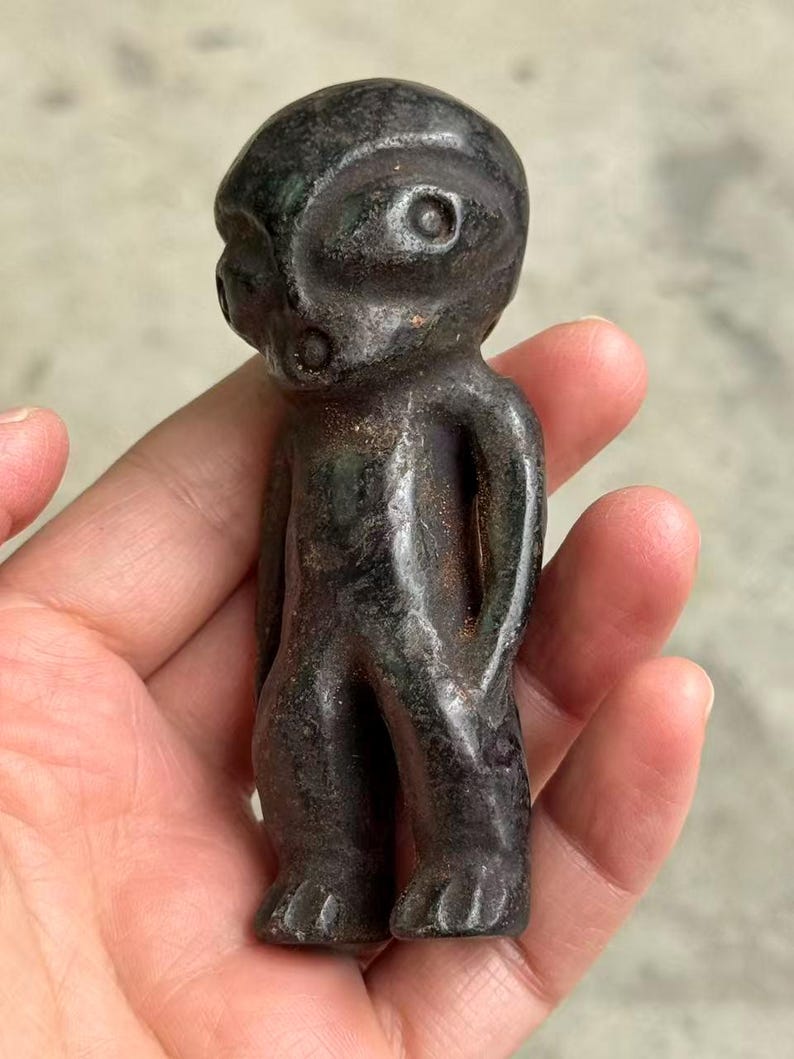 May include: A dark, weathered figurine of a humanoid figure with a large head and small body. The figure has a rounded helmet-like head with two eye-like indentations and a small mouth. The figure is held in a hand.