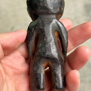 May include: A small, dark-colored figurine of a person with a rounded head and simple body. The figurine has a weathered appearance with hints of brown and green, suggesting age or a specific material. The figure is held in a hand.