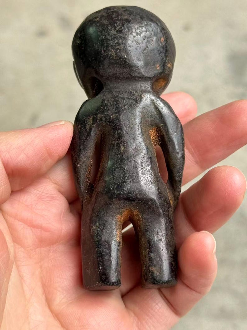May include: A small, dark-colored figurine of a person with a rounded head and simple body. The figurine has a weathered appearance with hints of brown and green, suggesting age or a specific material. The figure is held in a hand.