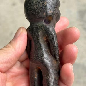 May include: A dark, weathered figurine of a small humanoid figure. The sculpture is a deep, mottled black with hints of green and purple. The figure has a rounded head and a simple, stylized form.