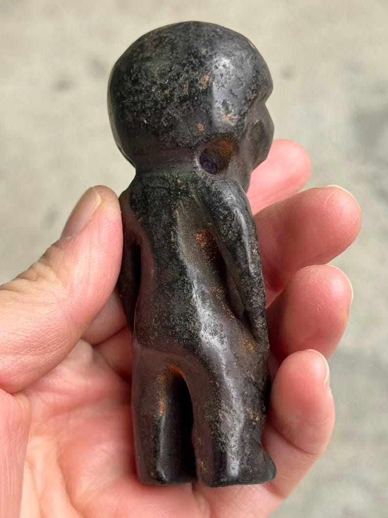 May include: A dark, weathered figurine of a small humanoid figure. The sculpture is a deep, mottled black with hints of green and purple. The figure has a rounded head and a simple, stylized form.