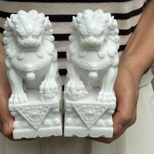 May include: Two white ceramic Foo Dog statues. Each statue is in a seated position, with detailed features including a mane, paws, and a decorative collar. The statues are mounted on square bases with triangular patterns.