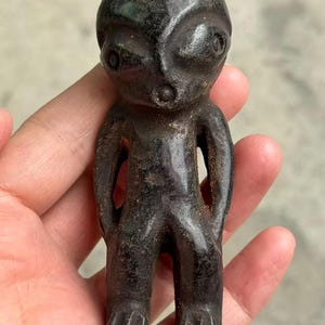 May include: A small, dark figurine of a humanoid form with large eyes and a rounded head. The figure has a simple, stylized form with arms at its sides and feet. The material appears to be a dark metal or stone, with a weathered appearance.