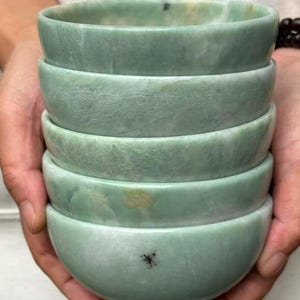 May include: Five light green stone bowls stacked together. The bowls have a smooth, rounded design with subtle colour and texture variations. The bowls are held in someone's hands.