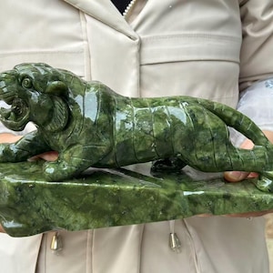 Op de afbeelding: Een gebeeldhouwde jade tijgersculptuur in groentinten. De tijger wordt afgebeeld in een hurkende positie met een open mond, rustend op een rechthoekige basis. De sculptuur wordt door een persoon vastgehouden.