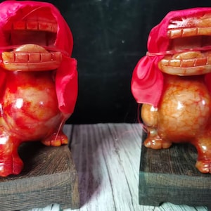 Può includere: Due statue di leone in pietra scolpita con colorazione screziata rossa e arancione. Ogni figura presenta una bocca aperta con denti visibili ed è drappeggiata con un panno di tessuto rosso brillante fissato da una sottile fascia rossa. Basi in legno.