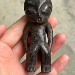 May include: A dark, weathered figurine of an alien-like humanoid figure. The small statue has a rounded head, large eyes, and a small mouth. The arms are at the sides, and the legs are together. The figure is held in a hand.