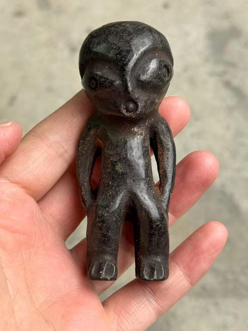 May include: A dark, weathered figurine of an alien-like humanoid figure. The small statue has a rounded head, large eyes, and a small mouth. The arms are at the sides, and the legs are together. The figure is held in a hand.