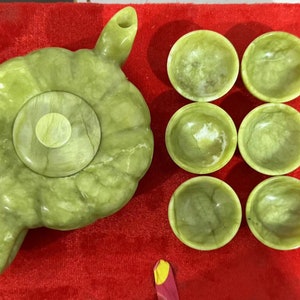 Op de afbeelding: Een lichtgroene jade theeset met een theepot en zes kleine kopjes. De theepot heeft een unieke, organische vorm met een handvat en tuit. De kopjes zijn rond en ondiep. De set wordt gepresenteerd op een rode ondergrond.
