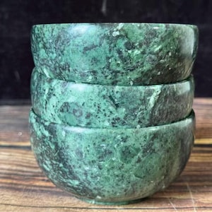 May include: A stack of three round bowls crafted from green stone with a mottled, dark-veined texture. Each bowl features a smooth, polished surface with varying shades of forest green and black, showcasing natural mineral patterns throughout the material.