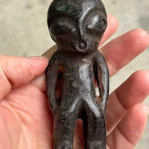 May include: A dark, weathered figurine of an alien-like figure. The small statue has a large head, large eyes, and a small mouth. The arms are at the sides, and the legs are together. The figurine is held in a hand.