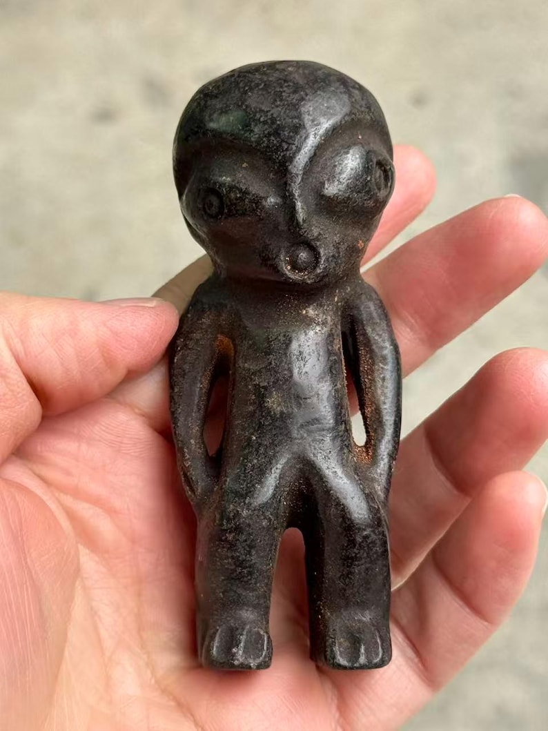 May include: A dark, weathered figurine of an alien-like figure. The small statue has a large head, large eyes, and a small mouth. The arms are at the sides, and the legs are together. The figurine is held in a hand.