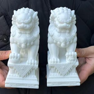 以下が含まれることがあります： 白いシーサーの像が2つ。各像は、詳細な鬣、顔の特徴、装飾的な襟を備えた、ライオンのような外観で精巧に彫刻されています。幾何学模様の長方形の台座の上にあり、伝統的なアジアの芸術様式を示しています。