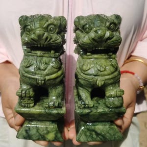 Può includere: Due statue di cani Foo verde giada. Ogni statua è scolpita nei dettagli, raffigurante un leone guardiano con un'espressione feroce. Le statue sono su basi quadrate e hanno una finitura lucida. I cani Foo sono un simbolo di protezione e prosperità.