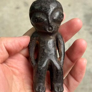 May include: A dark, weathered figurine of an alien-like humanoid figure. The sculpture has a rounded head, large eyes, and a small mouth. The arms are at the sides, and the legs are together. The figure is held in a hand.