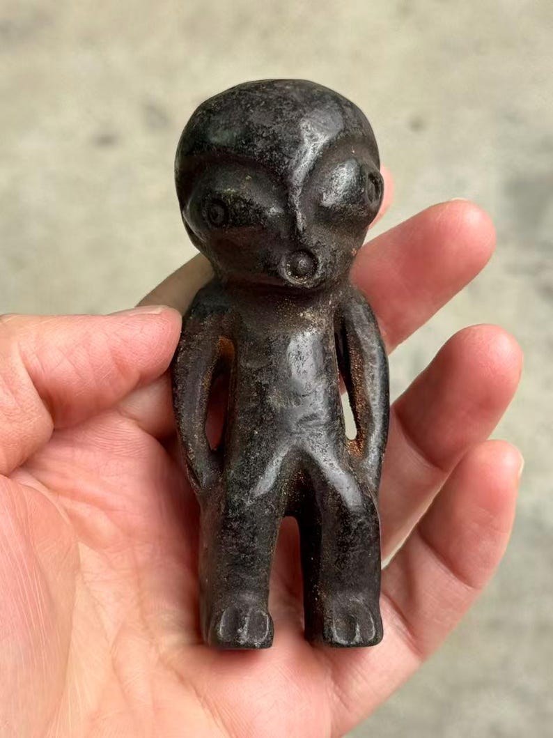 May include: A dark, weathered figurine of an alien-like humanoid figure. The sculpture has a rounded head, large eyes, and a small mouth. The arms are at the sides, and the legs are together. The figure is held in a hand.