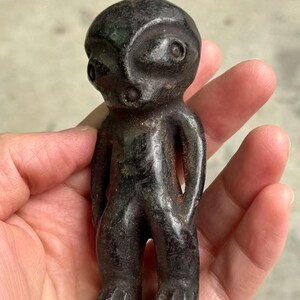May include: A small, dark-colored figurine of a humanoid figure. The figure has a rounded head, large eyes, and a simple body with arms at its sides. The figurine is held in a hand.
